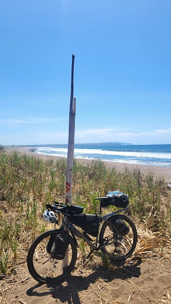 Ein Fahrrad mit Gepäck steht am Strand im Sand; dahinter Meer, Wellen und blauer Himmel.