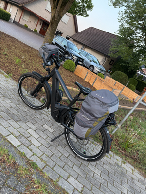 Ein schwarzes, voll beladenes Tourenrad steht auf einem Gehweg; dahinter Wohnhäuser, parkende Autos und Bäume.